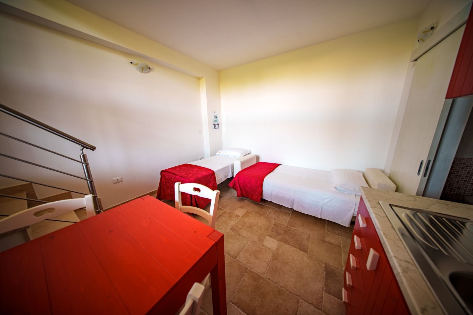 Due letti singoli con coperte rosse in una stanza semplice, piccola cucina, tavolo rosso e scale.