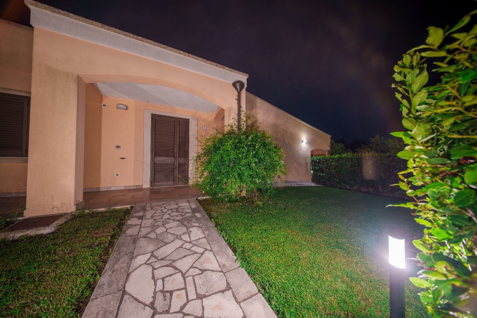 Esterno di una casa beige di notte, sentiero in pietra che conduce all'ingresso, con giardino illuminato.