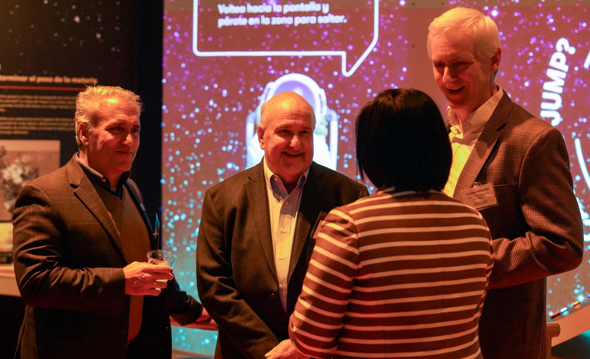 Four people talking at an exhibit. Dark suits and sweaters. Background includes display with text and image.