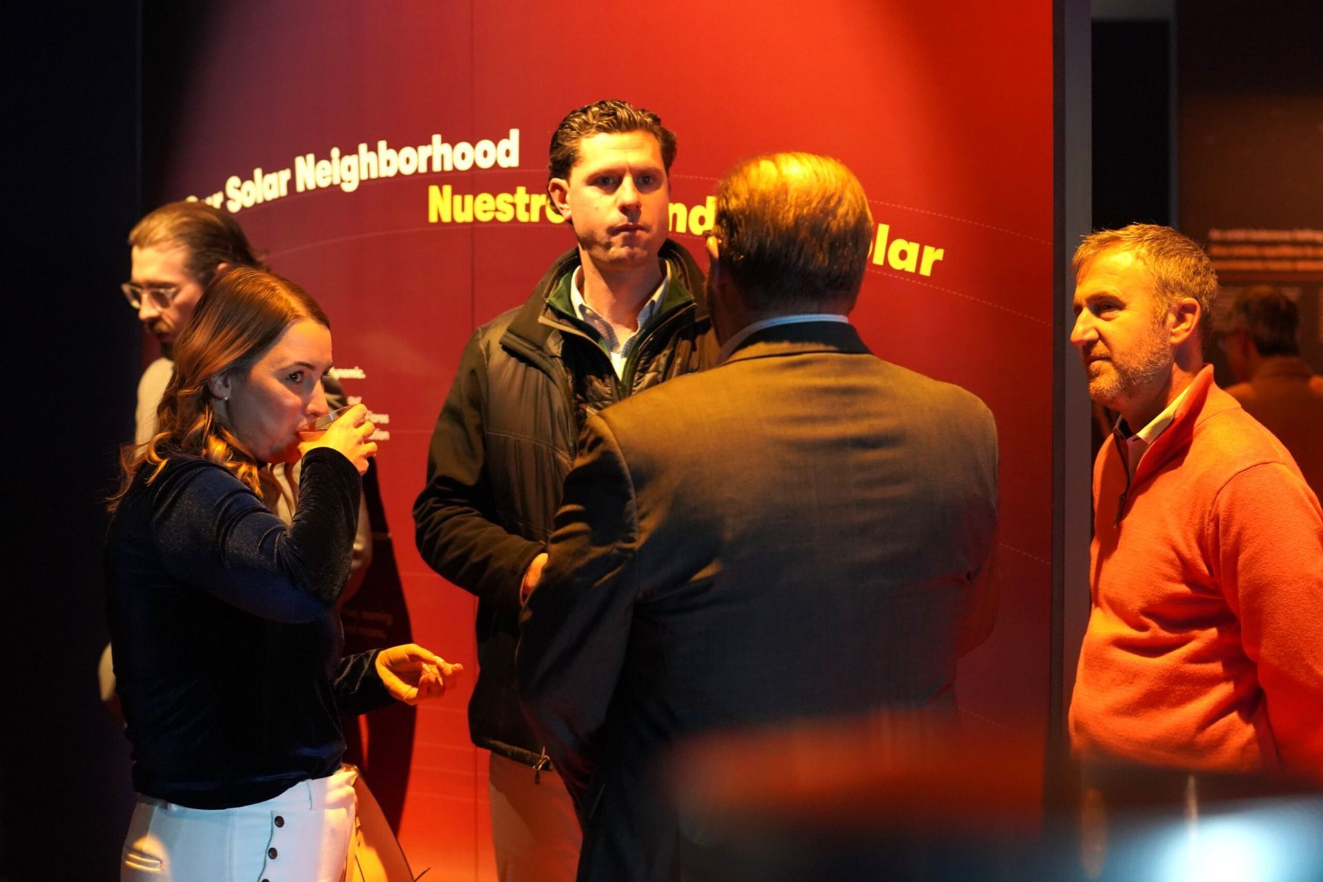 Five people gathered near a red wall with text. A woman drinks from a cup; others converse.