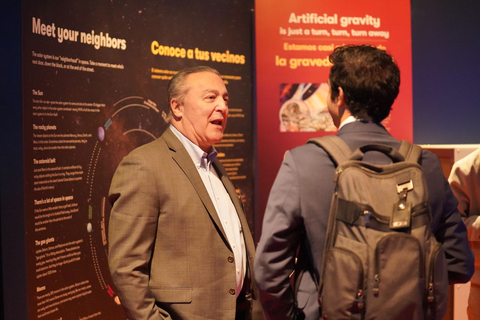 Man in blazer speaks to another man with a backpack near exhibit panels about space.