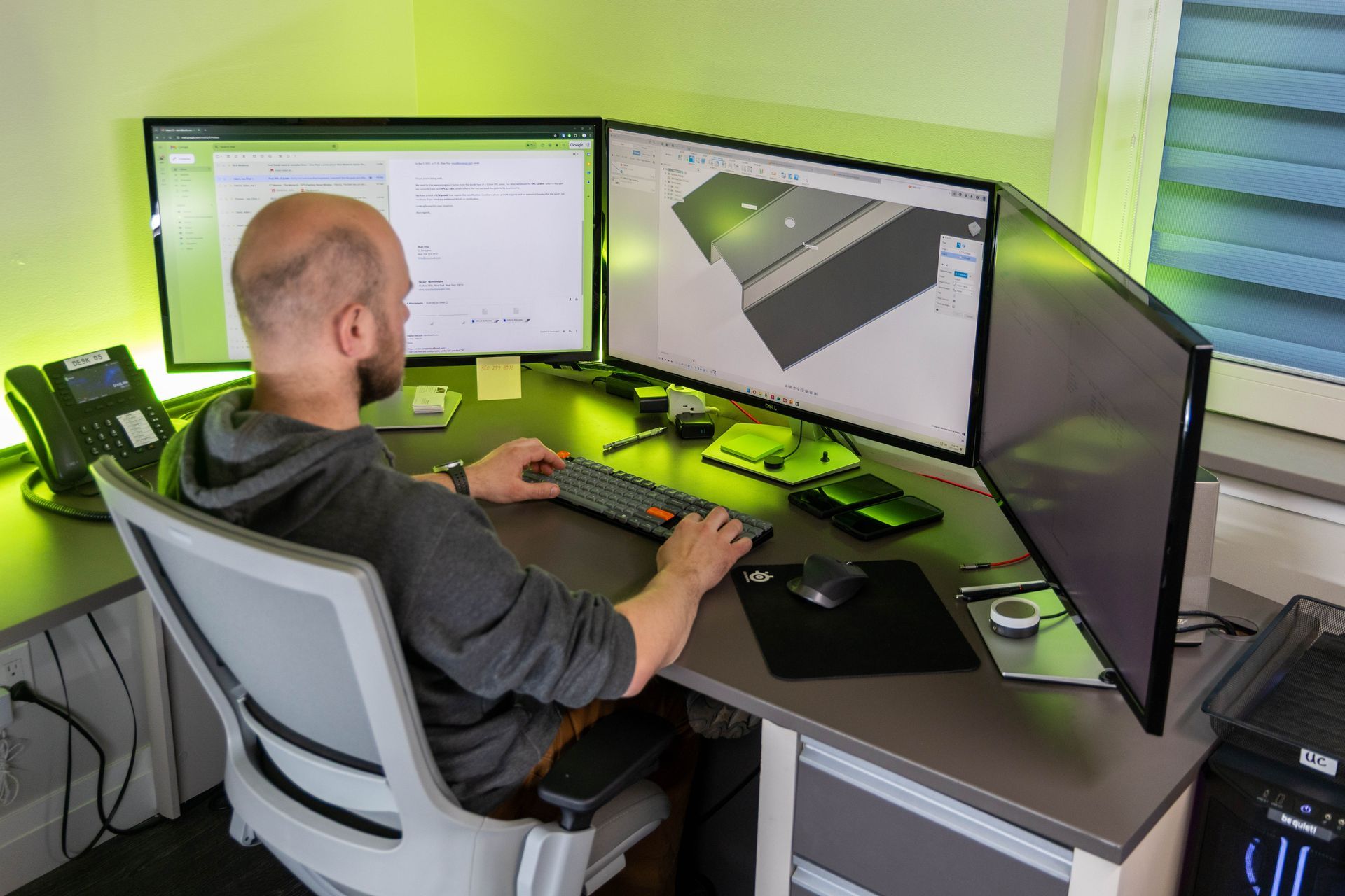 Man working at a desk with three monitors, a keyboard, and a mouse; lit by green light.