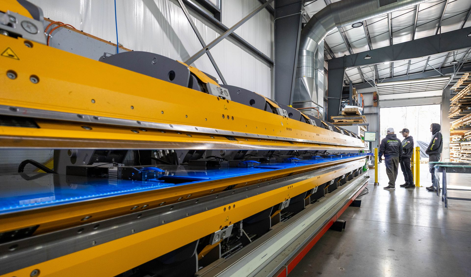 Large industrial machine, yellow and blue, in a factory setting. Three workers observe.