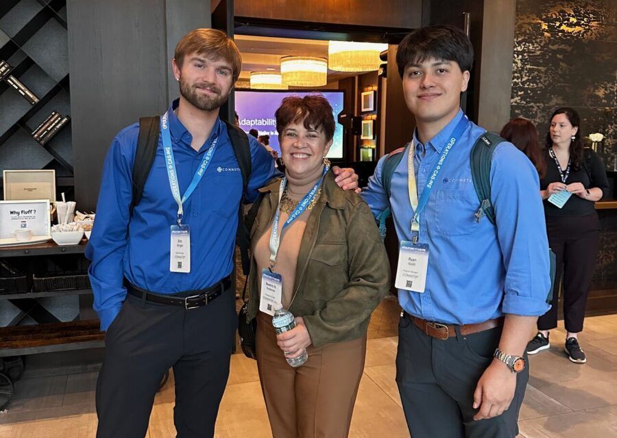 Three people standing indoors; two men in blue shirts flank a woman in brown. All wear lanyards, smiles.
