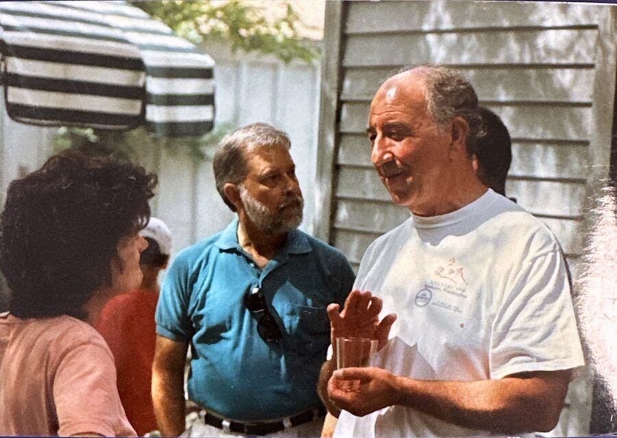 Three men in an outdoor setting talking. One gestures with a glass. Light-colored shirts, a striped awning.