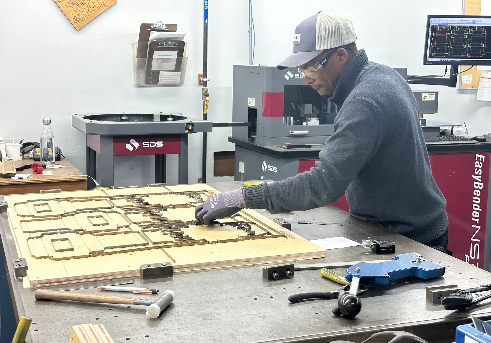 Man in a workshop inspecting a wood die, with machinery in the background. He wears gloves and a cap.