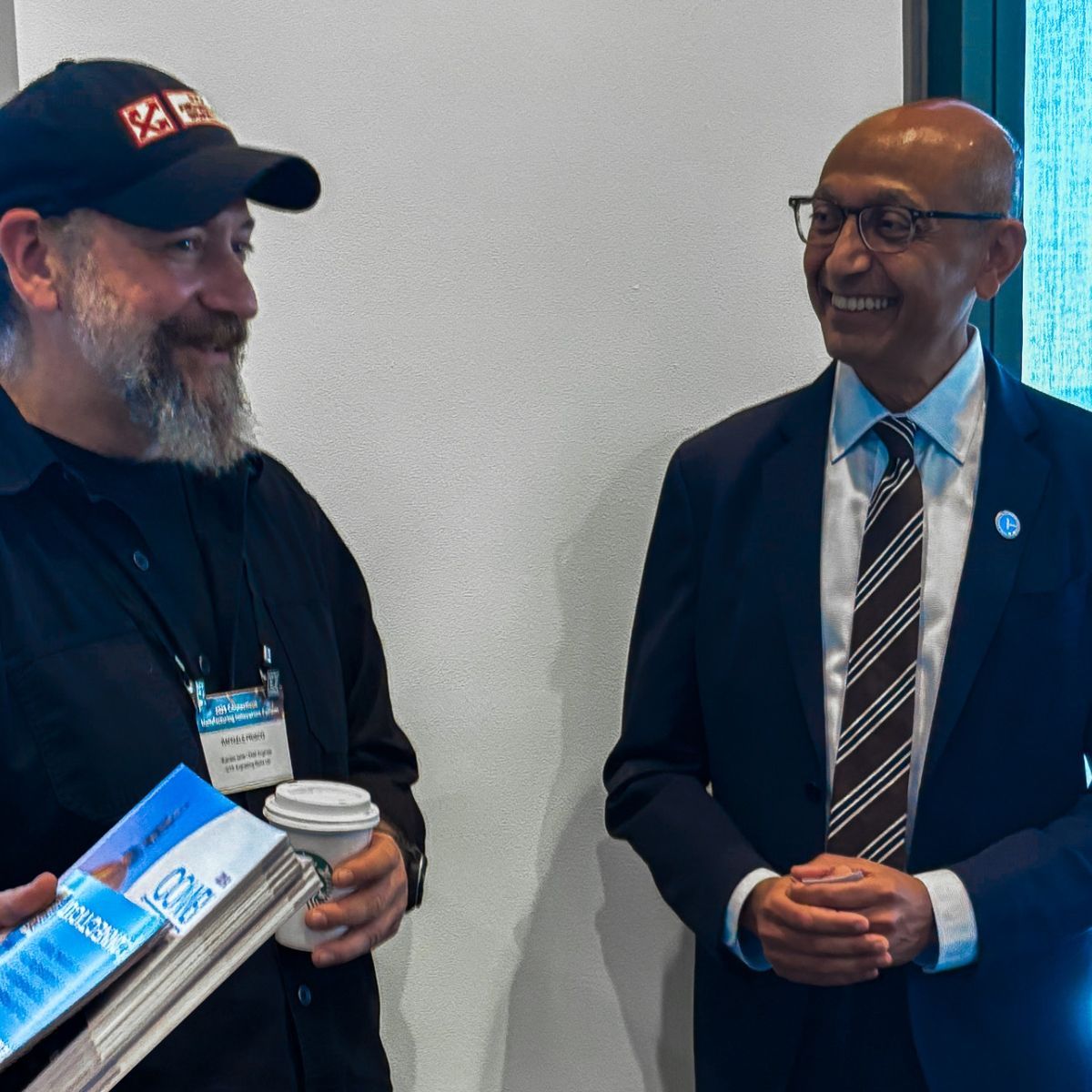 Two men smiling at each other. One wears a black cap and shirt, holding books and a coffee. The other wears a suit and tie.