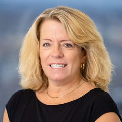 A portrait of Kelly Tubridy, Chief Executive Administrator of CONNSTEP, smiling, wearing a black top and gold jewelry, against a blurred background.
