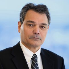 A portrait of Matin Karbassioon, Continuous Improvement at CONNSTEP, wearing a dark suit, looking at the camera with a neutral expression. Blurred blue background.
