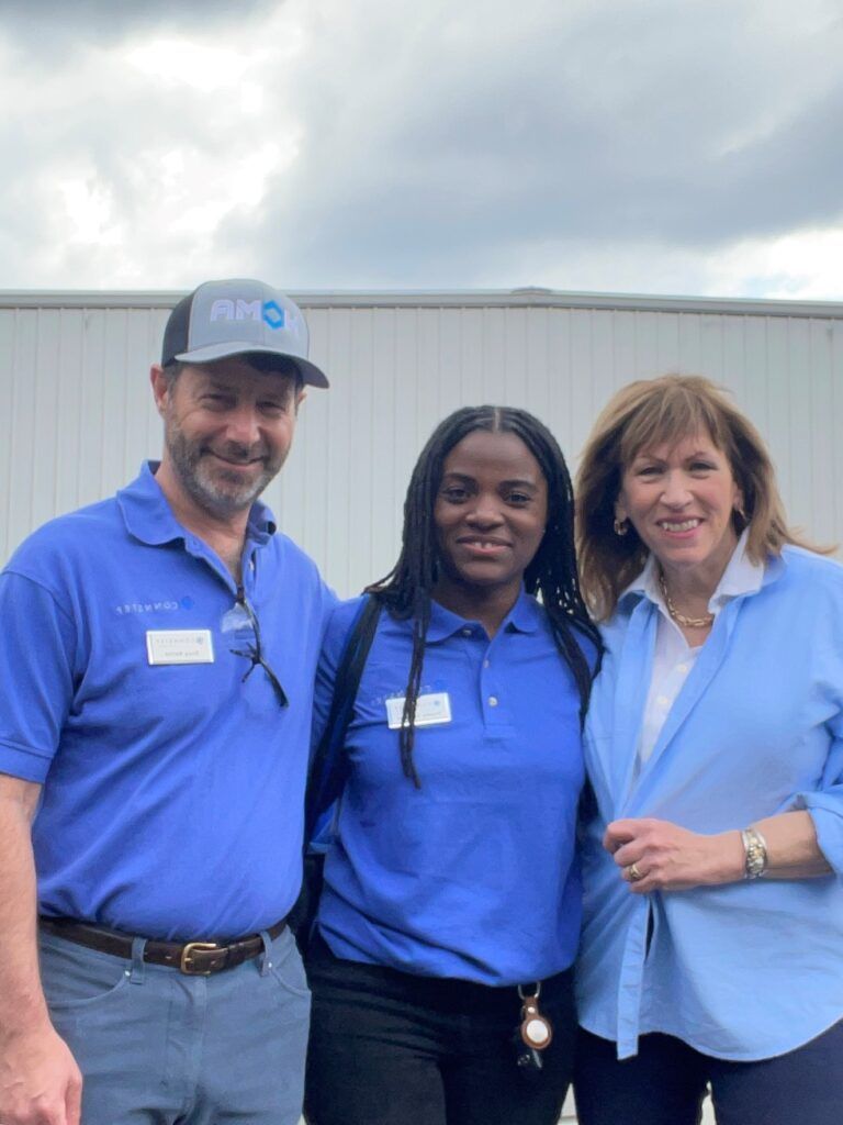 Three people in blue shirts stand together, smiling in front of a white building.
