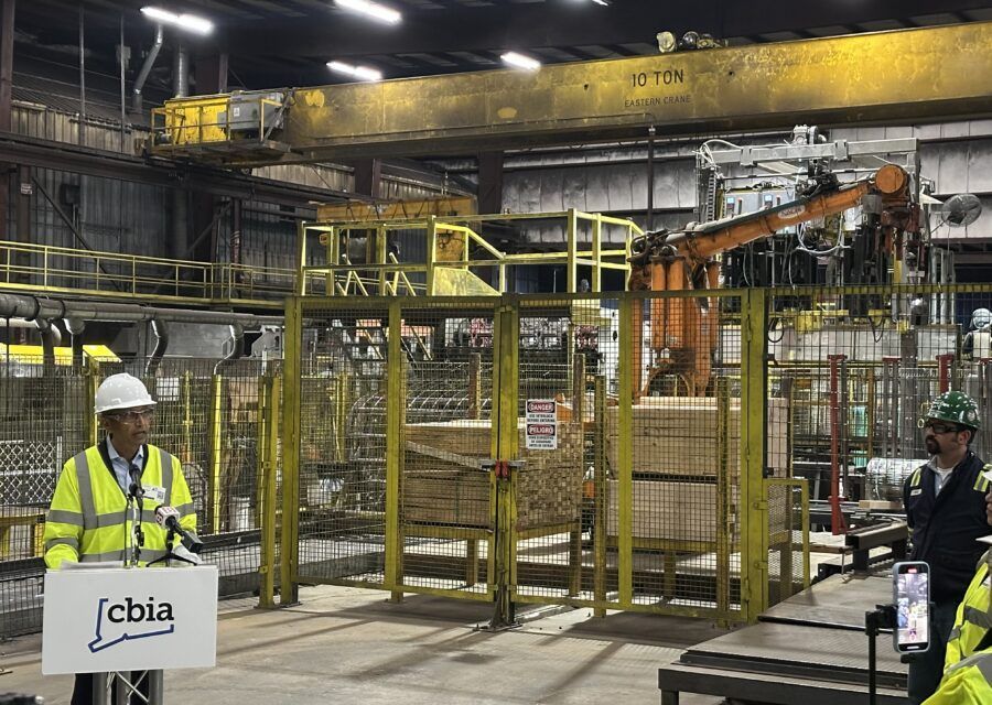 Man at a podium with microphone; factory setting with industrial robot arm, stacks of wood, and machinery.