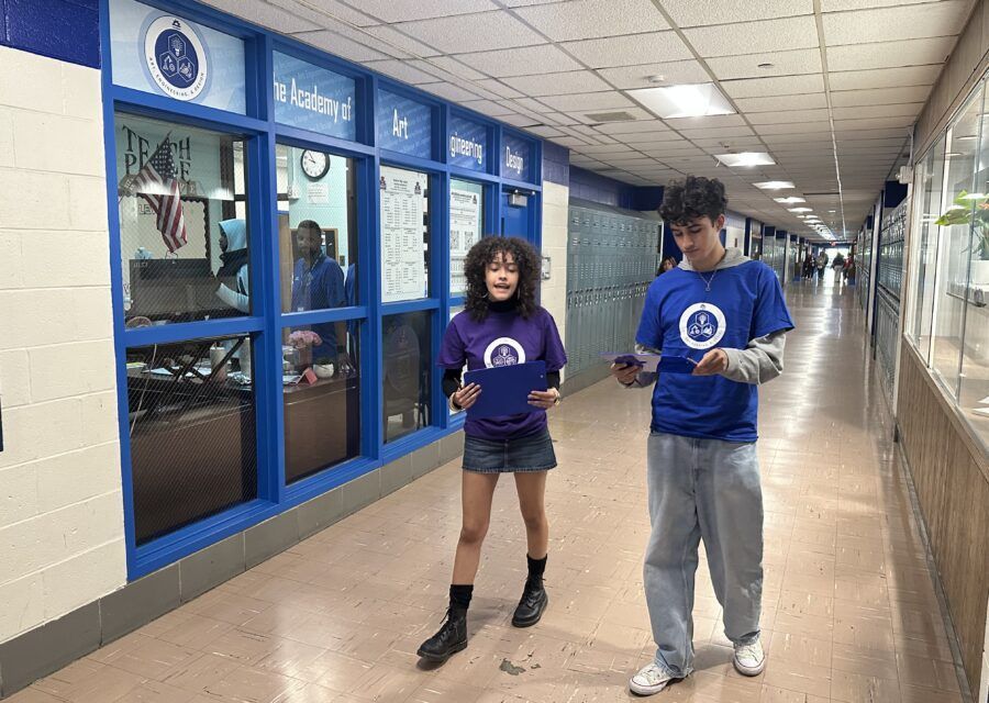 Two people in matching blue shirts walk down a hallway, holding papers. Blue and white walls and windows.