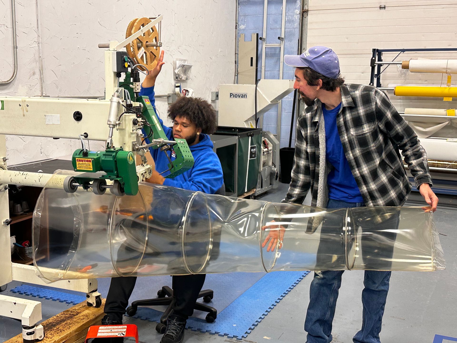 Two people working with clear plastic on a machine in a workshop. One holds a roll, the other guides the plastic.