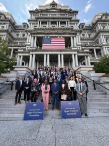 Group of people posing on steps in front of a large building with a US flag. Blue signs in front.