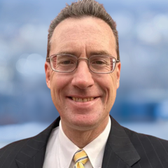 A portrait of Brian Burnim, Business Development Advisor at CONNSTEP, with short, light brown hair smiling, wearing a white shirt, yellow tie, black suit jacket with a blurred blue and white background.