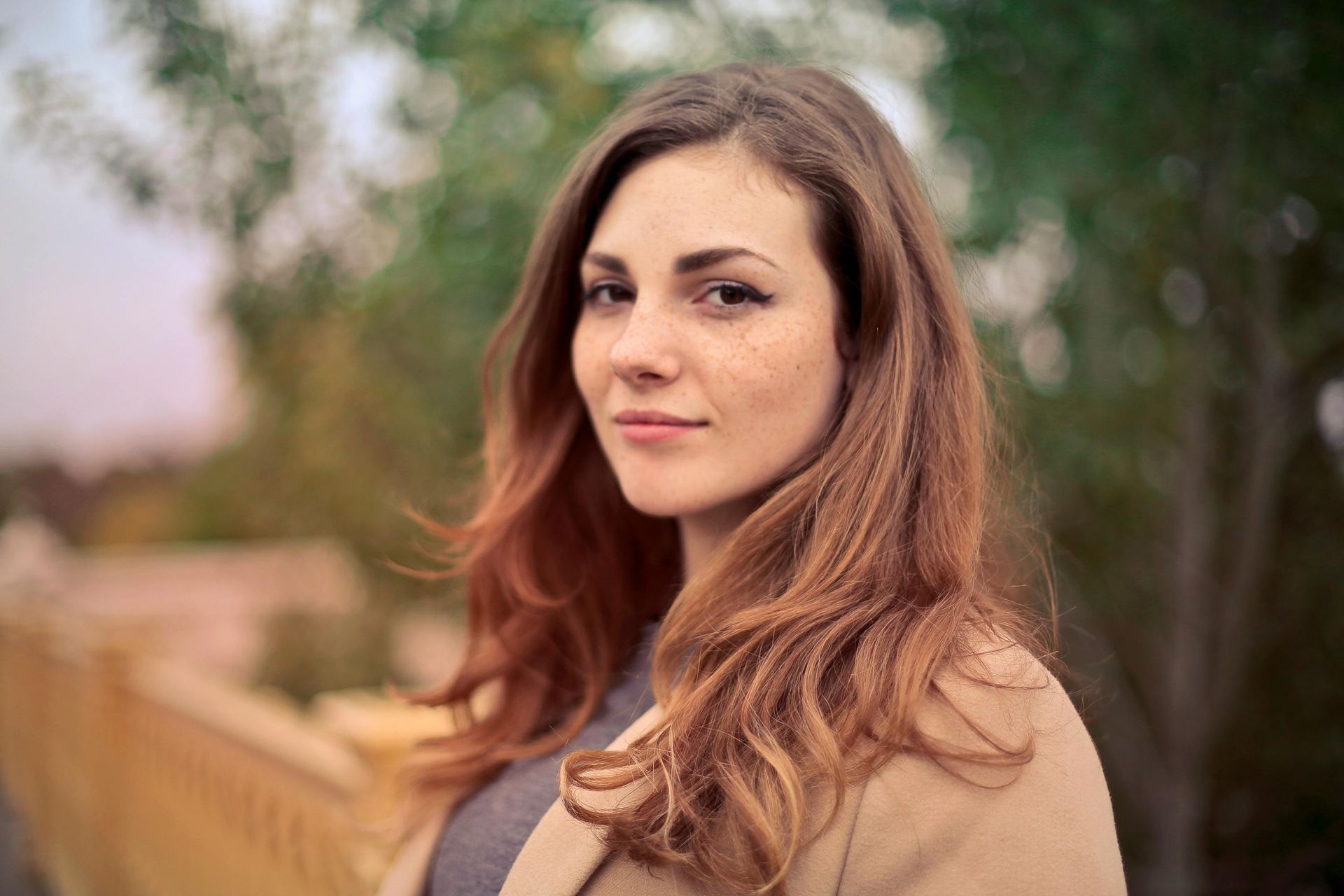 Woman with auburn hair and freckles, looking at the camera, wearing a light-colored coat, outdoors.