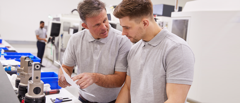 Two men in a manufacturing facility looking at papers. One man is pointing.