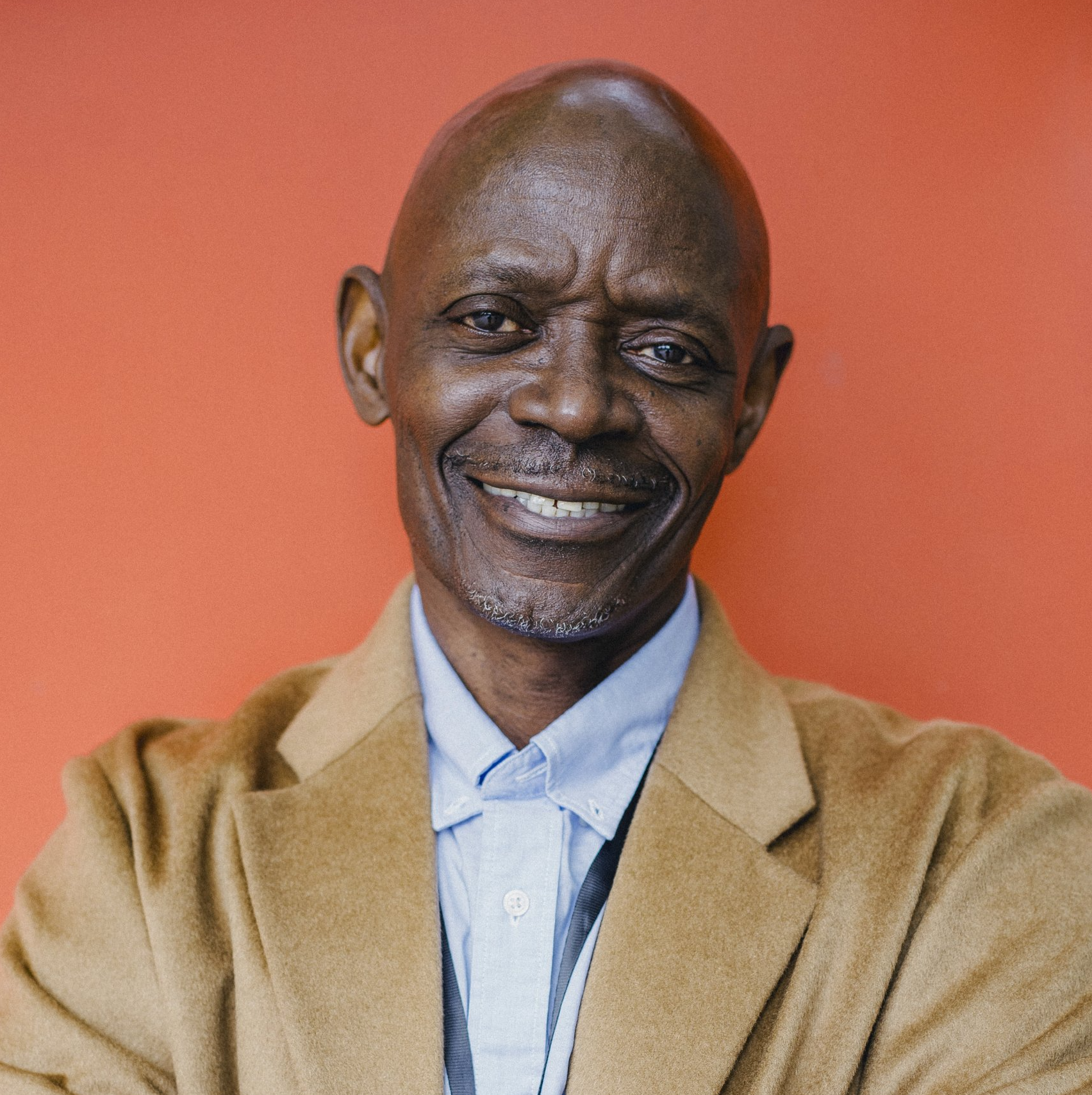 A man in a tan jacket and blue shirt is smiling with his arms crossed