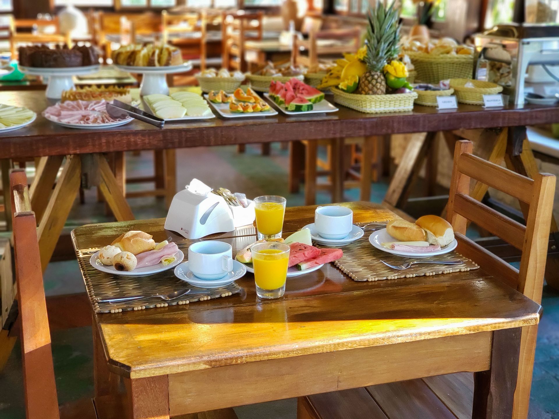 Mesa de madeira posta para o café da manhã com café, suco de laranja, doces e um buffet de frutas ao fundo.