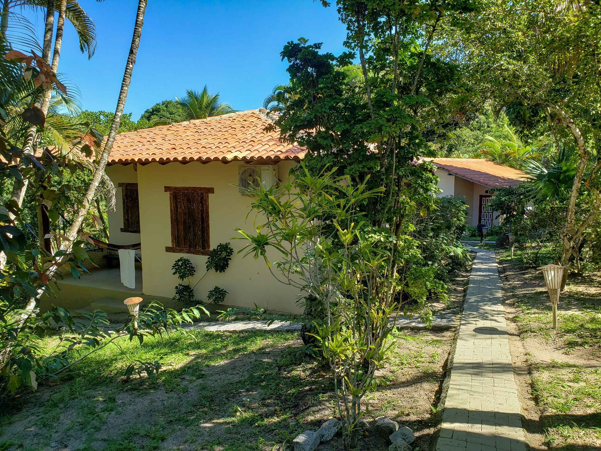 Casa tropical com telhado de telha vermelha e caminho de jardim em meio a árvores e plantas verdejantes.