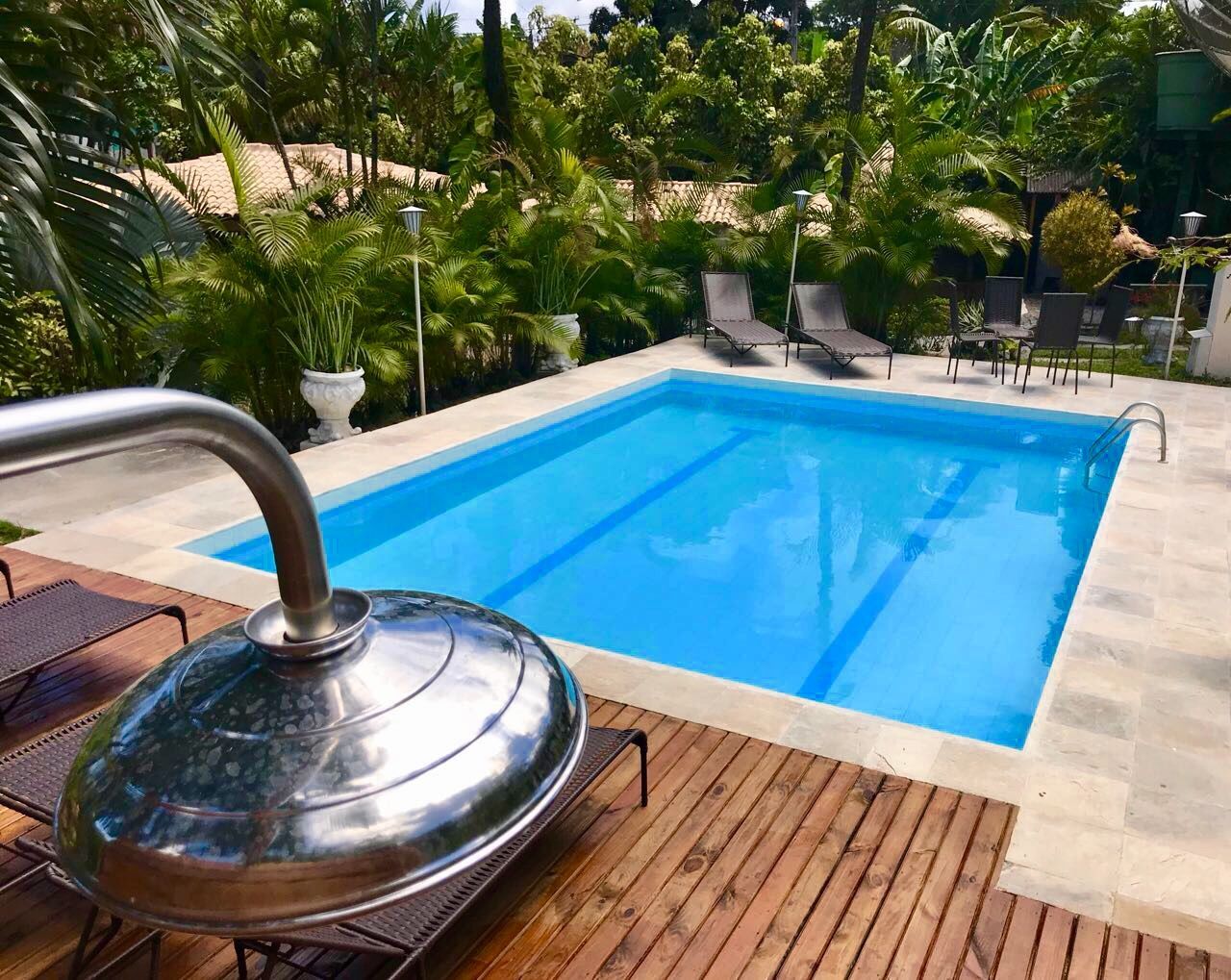 Piscina no quintal com água azul, espreguiçadeiras, plantas tropicais e um chuveiro de aço inoxidável em primeiro plano.