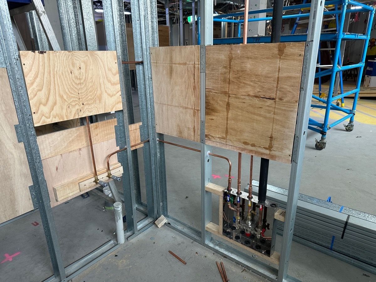 Photograph of an indoor construction site showing exposed metal wall studs with plumbing pipes installed within the framing.