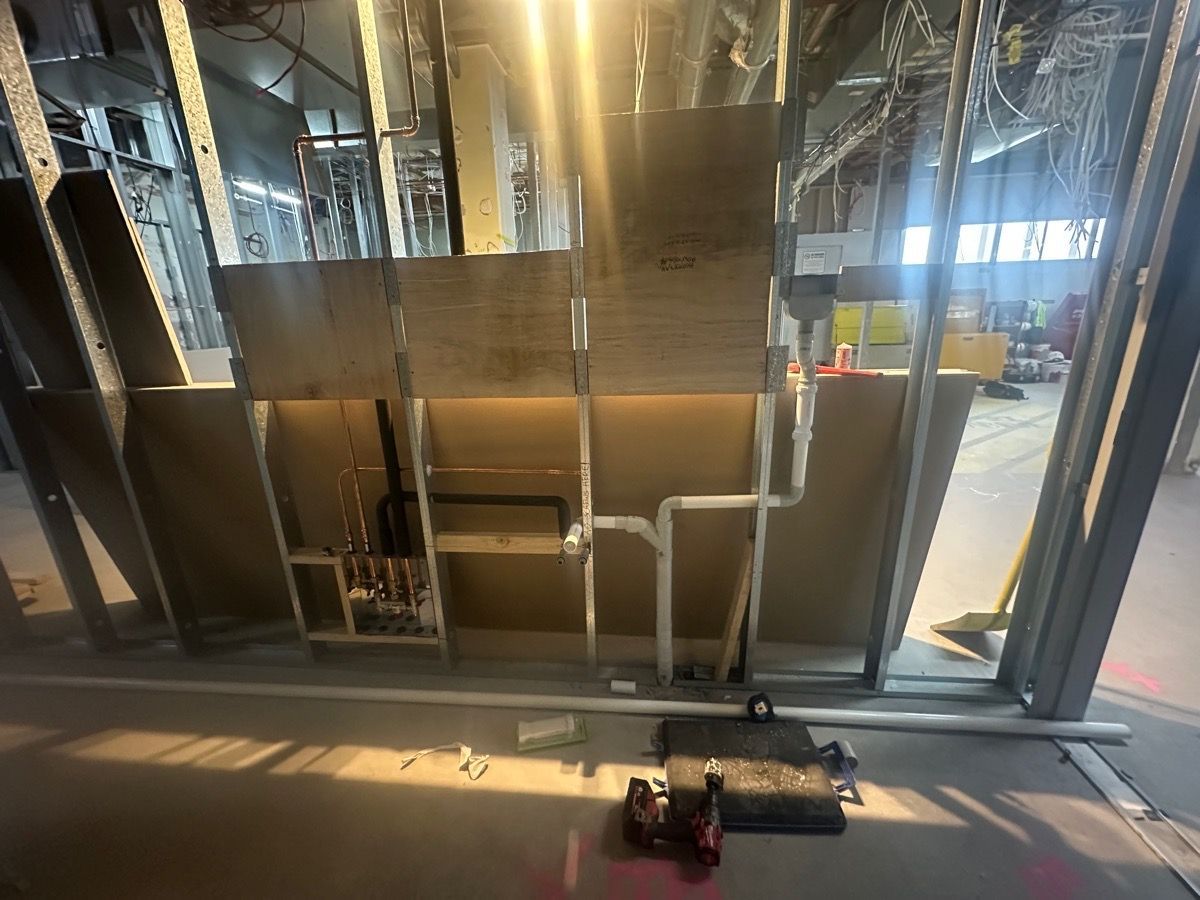 Photograph of an indoor construction site showing exposed metal wall studs with plumbing pipes installed within the framing.