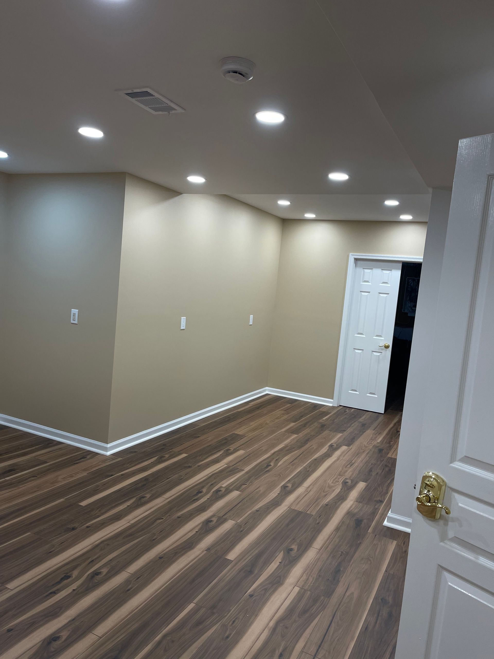 Empty beige room with wood-look flooring, recessed ceiling lights, and an open white door.
