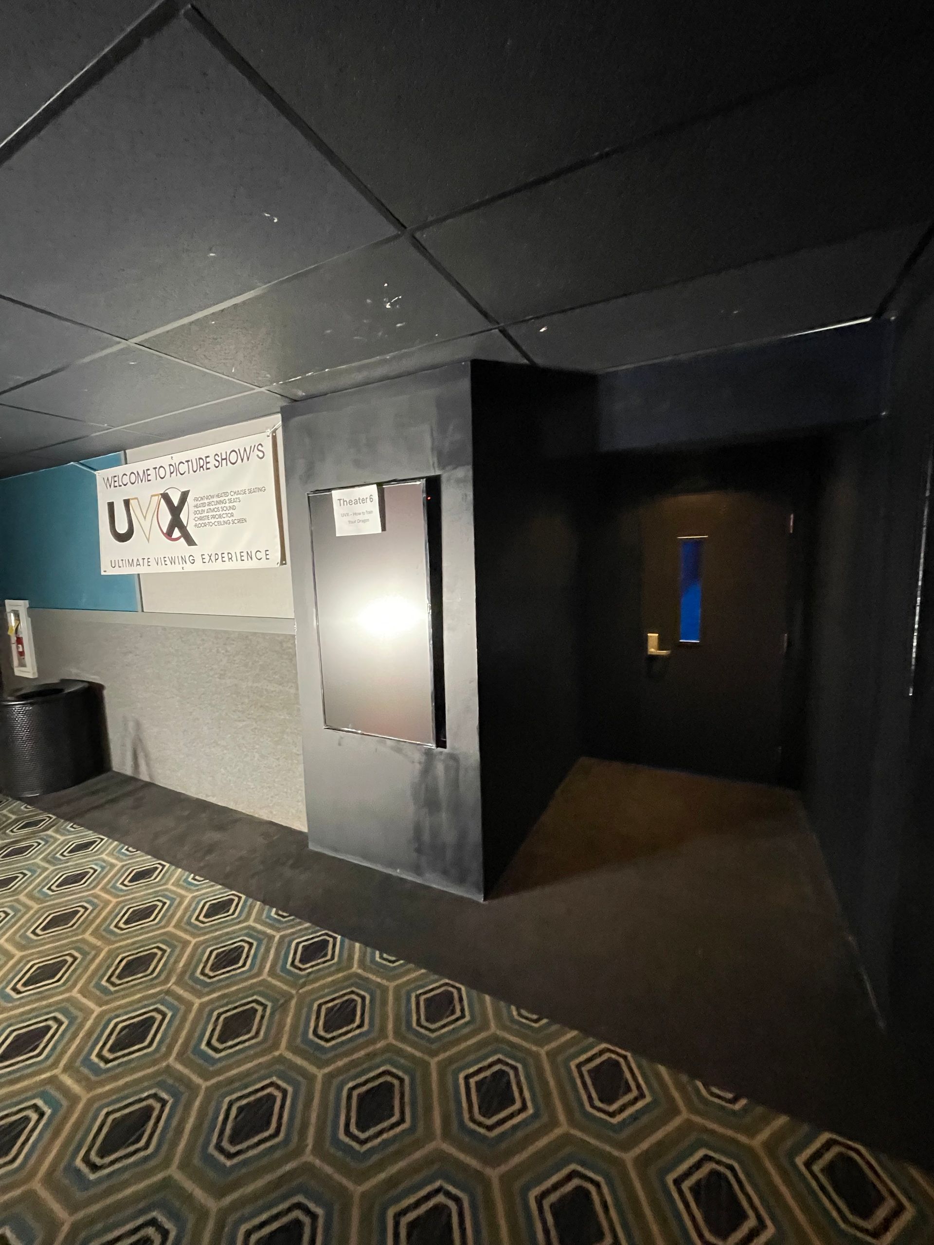 Dim hallway with patterned carpet, dark walls, a black door, and a poster on the left wall.