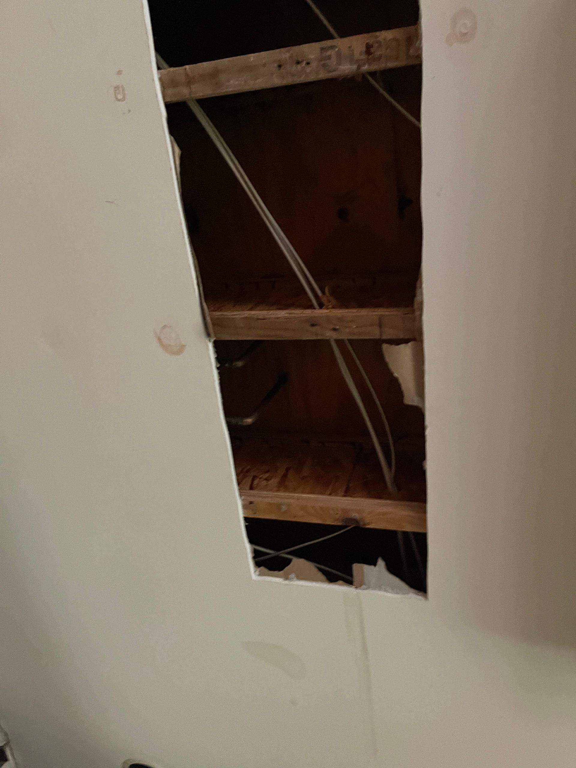 Damaged white ceiling with a rectangular hole exposing wooden beams and wiring