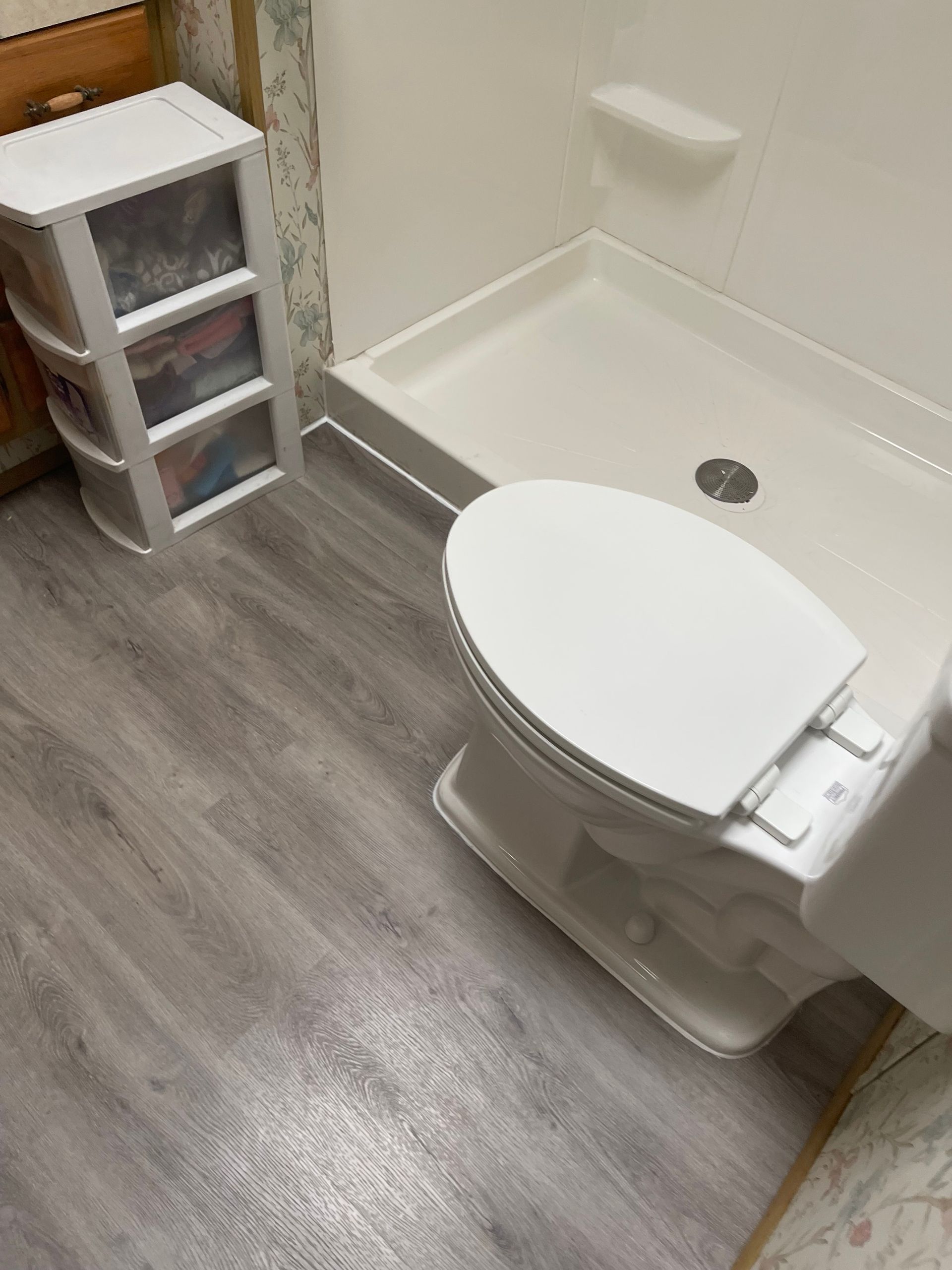 Small bathroom with white toilet, shower tub, and plastic drawer unit on gray wood-look floor.