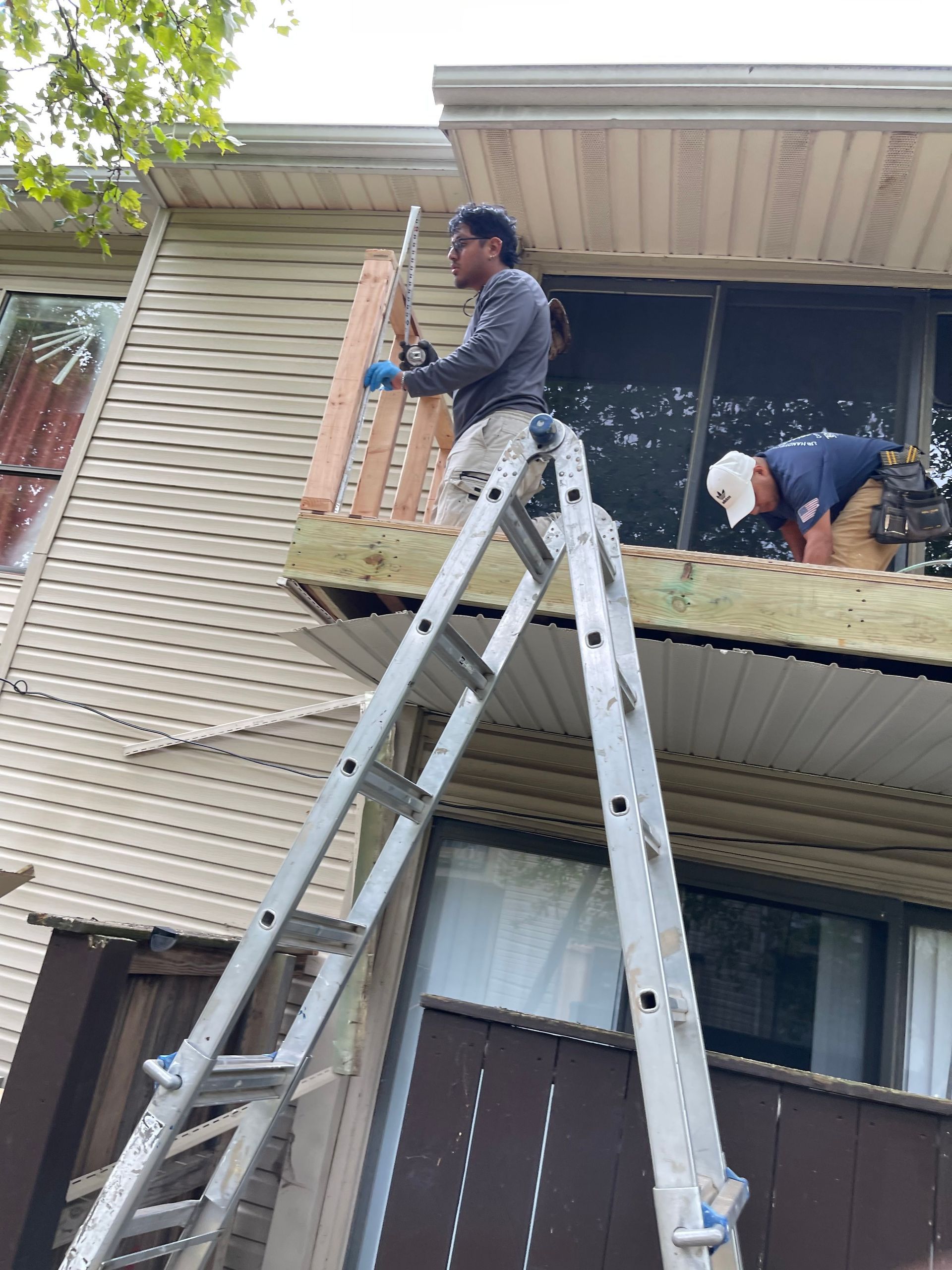 Two workers on ladders repairing an apartment balcony under a corrugated roof