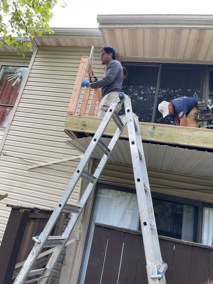 Two workers on ladders repair an apartment balcony and exterior siding.