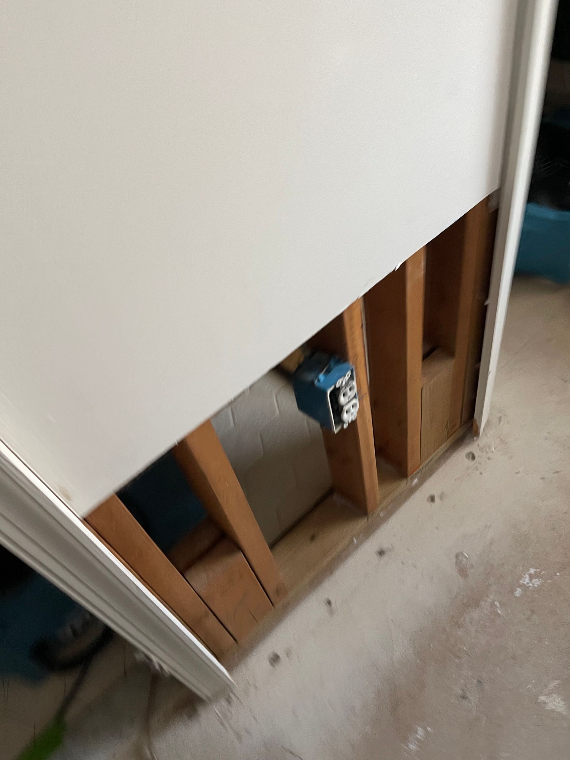 Open wall with exposed wooden studs and a blue electrical box near a door frame on a concrete floor