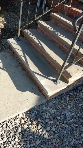 Small concrete front steps with black handrails leading up to a doorway, next to gravel and sunlight.