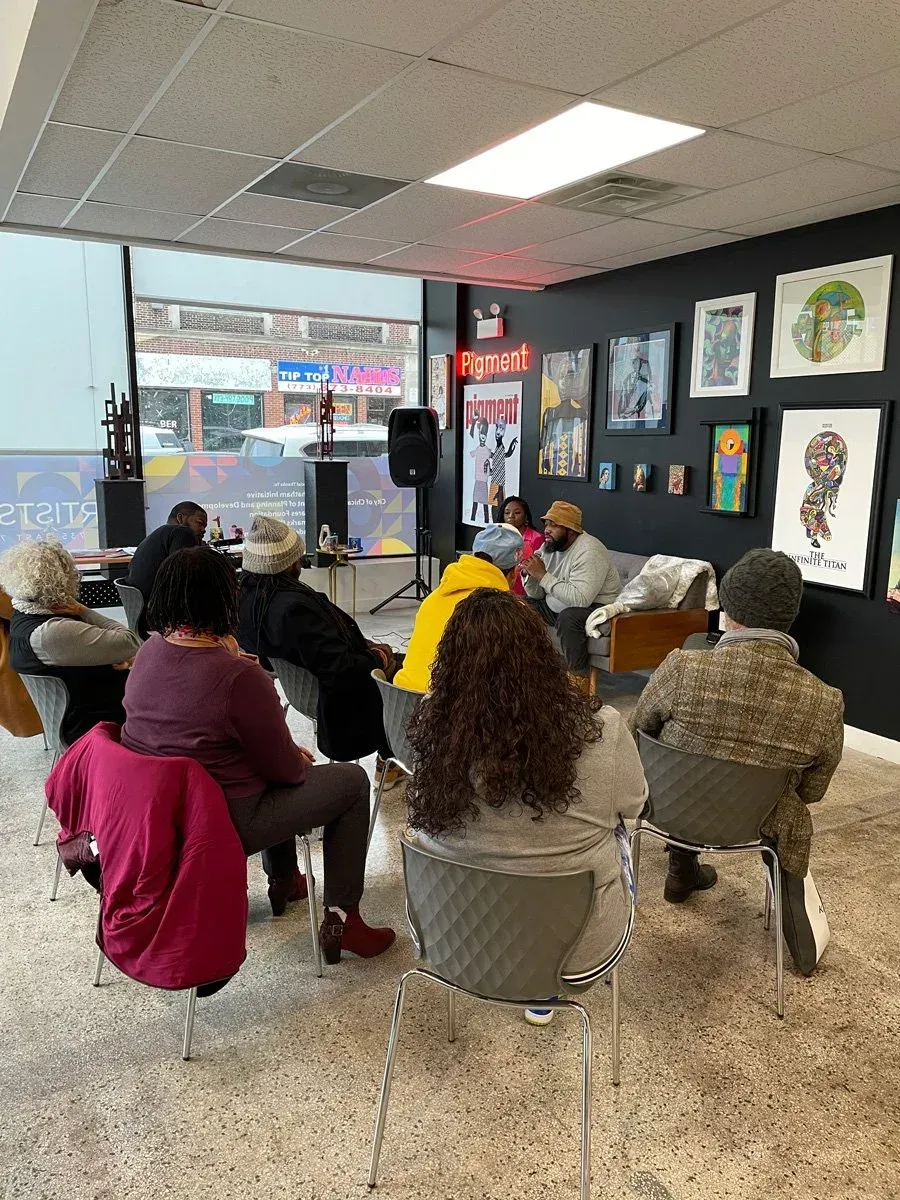 People seated in a small gallery or shop, listening near a front window with framed artwork on the walls.