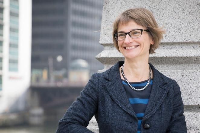 Woman smiling, wearing glasses and a blazer, outdoors near a concrete structure, city background.
