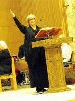 Woman speaking at a podium, gesturing with one arm raised in a wood-paneled room
