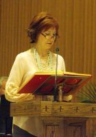 Person speaking at a lectern, reading from a red folder in a wood-paneled room.