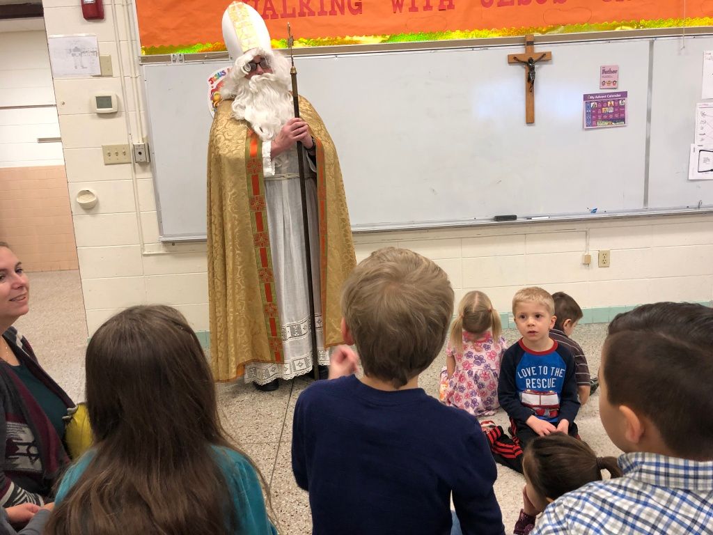 Teacher in a white robe stands before seated children in a classroom with a whiteboard and cross.