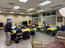 People seated around yellow-covered tables in a community room, with two standing near the front.