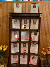 Memorial shelf with framed photos and lit candles, flanked by flower arrangements against a wood-paneled wall