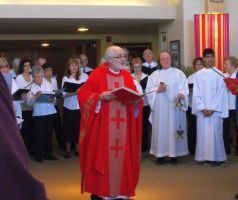 Catholic priest in red vestments reading from a book before a choir in a church hall