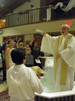 Priest in white robes baptizes a person in a church baptismal font while onlookers watch.