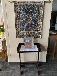 Small table beneath a patterned wall hanging, with a framed photo and white lace runner in a hallway