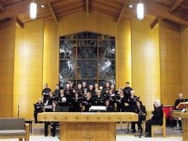 Choir performing in a wood-paneled church, with a lit altar and stained-glass window behind them.
