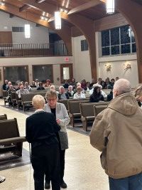 People seated in a church or community hall, with three people standing in the foreground.