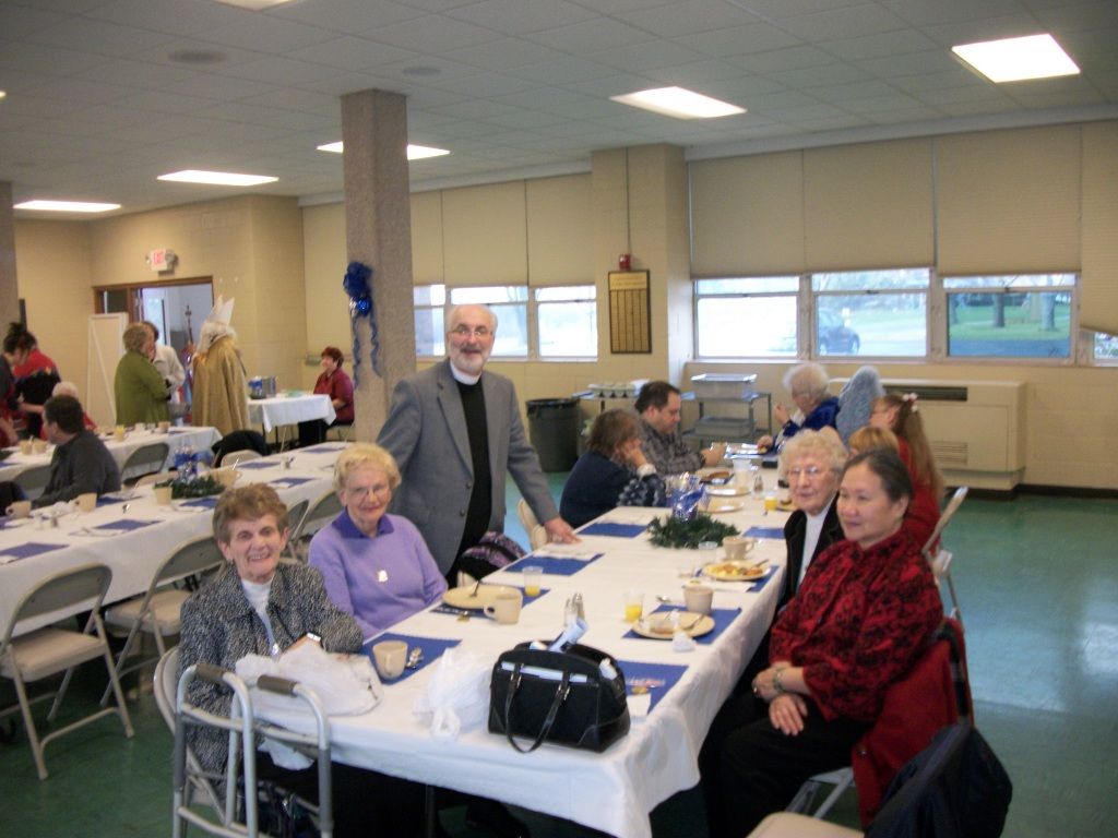 People seated at long tables in a community hall, eating and talking during a social gathering.