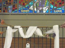 Church altar with white draped cloth and stained glass reading “Saint Luke” above a metal railing