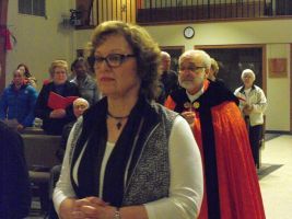 People standing in a hall, with a woman in glasses and a red-robed man in front.